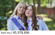Two female graduates with purple sashes and bow ties embrace with closed eyes, commemorating their last school day. Стоковое фото, фотограф Mikhail Davidovich / Фотобанк Лори