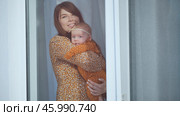 Smiling mother holding her baby girl by the window at home, both wearing matching dresses. Стоковое фото, фотограф Mikhail Davidovich / Фотобанк Лори