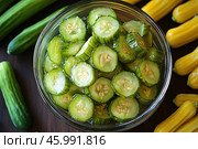 Fresh cucumbers sliced ​​into circles in a glass bowl. The cucumbers look juicy and bright green, with a characteristic shine indicating freshness. Whole cucumbers and yellow mini-zucchinis lie around the bowl, creating a color contrast and adding naturalness to the composition. The background is done in warm tones, which emphasizes the naturalness and appetizing nature of the image. Стоковое AI-изображение, AI-художник Анна Кривицкая / Фотобанк Лори