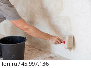 A hand with a wide brush and a bucket next to the wall being repaired during the application of primer or glue before gluing wallpaper. Wall treatment with a primer. Стоковое фото, фотограф Photojuli / Фотобанк Лори