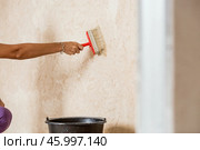 A hand with a wide brush and a bucket next to the wall being repaired during the application of primer or glue before gluing wallpaper. Wall treatment with a primer. Стоковое фото, фотограф Photojuli / Фотобанк Лори