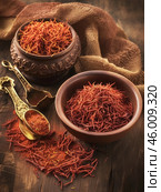 Saffron threads scattered and placed in two bowls: one of them is made of ceramic with a carved pattern, the other is of a simpler shape. Also visible is an elegant metal spoon with a decorative handle, on which lies a little saffron. The background is made of textured wood, creating a feeling of warmth and coziness, and around it is laid out a coarsely knitted fabric, enhancing the atmosphere of naturalness and tradition. Стоковое AI-изображение, AI-художник Анна Кривицкая / Фотобанк Лори