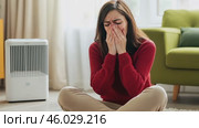 The woman sneezes her face in her hands at home. Sick young woman blowing her nose. Female sneezing sitting on the floor with heater. Allergy, cold or flu. Hay fever, viral infection, health problem. Стоковое AI-видео, аниматор Марина Демидюк / Фотобанк Лори