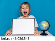 Funny little preschool girl with open mouth, sitting at table with empty white mock up paper board and a globe on a light blue background, copy space. Preschool education, traveling with children. Стоковое фото, фотограф Ирина Михайличенко / Фотобанк Лори