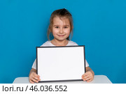 Happy little preschool girl sitting at table with empty white mock up paper board on a light blue background, copy space. Preschool education, goods for children. High quality photo. Стоковое фото, фотограф Ирина Михайличенко / Фотобанк Лори
