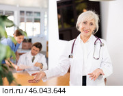 positive senior medical female colleague standing in meeting room and employees are talking behind her at table. Стоковое фото, фотограф Яков Филимонов / Фотобанк Лори