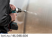 A man holds a battery-powered screwdriver and assembles a new greenhouse made of polyethylene, polycarbonate. Macro photography. Side view. Preparing for the spring garden season. Стоковое фото, фотограф Photojuli / Фотобанк Лори