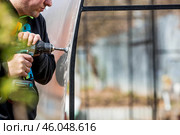 A man holds a battery-powered screwdriver and assembles a new greenhouse made of polyethylene, polycarbonate. Macro photography. Side view. Preparing for the spring garden season. Стоковое фото, фотограф Photojuli / Фотобанк Лори