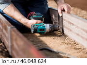A man holds a battery-powered screwdriver and collects new beds from WPC into a greenhouse made of polyethylene, polycarbonate. Macro photography. Side view. Preparing for the spring garden season. Стоковое фото, фотограф Photojuli / Фотобанк Лори