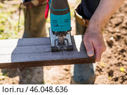 A man holds a battery-powered electric saw and builds new beds in a greenhouse made of wood-polymer composite. Macro photography. Side view. Preparing for the spring garden season. Стоковое фото, фотограф Photojuli / Фотобанк Лори