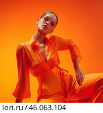 Stylish young woman in an elegant orange outfit consisting of a low-cut shirt and high-waisted trousers tied with a belt. She poses confidently and gracefully, slightly tilting her head, which adds dynamics to the image. The background is made in gradient shades of orange, creating the effect of unity with the model's clothes. The image is complemented by glasses with a thin frame and exquisite earrings, giving a modern and fashionable look. Стоковое AI-изображение, AI-художник Анна Кривицкая / Фотобанк Лори