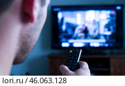 Man watching television with remote control in hand, close-up view of person holding TV remote, blurred screen in background, relaxing at home, leisure activity, focus on remote. Стоковое AI-изображение, AI-художник Павел Муравьев / Фотобанк Лори