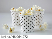 Popcorn overflowing from dotted paper cups on a white table is ready for a movie night. Стоковое AI-изображение, AI-художник Иван Рябоконь / Фотобанк Лори