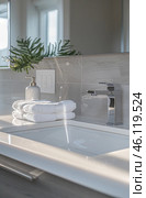 Modern bathroom with minimalist and stylish design. White washbasin with smooth lines is built into the glossy countertop. Above the washbasin is an elegant chromed metal mixer. Three snow-white towels are neatly folded nearby, creating a feeling of cleanliness and freshness. Behind you can see a soap dispenser in a white ceramic body with a golden dispenser, and next to it is a socket with switches. In the background there is a decorative plant, giving the interior a natural touch. Light comes through the window, creating soft shadows and emphasizing the harmony of the space. Стоковое AI-изображение, AI-художник Анна Кривицкая / Фотобанк Лори