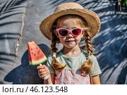 A little girl in sunglasses eats a watermelon in the hot summer. Funny happy child with a watermelon in his hands. Baby looks at the camera, healthy food. Стоковое AI-изображение, AI-художник Photojuli / Фотобанк Лори