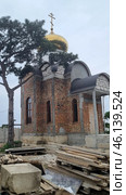 An Orthodox church under construction with a golden dome and an Orthodox cross on top. The walls of the building are made of red brick, in some places not yet covered with a finishing layer. The architectural style is reminiscent of traditional Russian churches, with characteristic semicircular arches above the windows and entrance. Стоковое фото, фотограф Анна Кривицкая / Фотобанк Лори