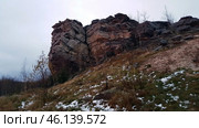 A rock with a characteristic layered structure surrounded by autumn nature. In the foreground, bushes with yellowed leaves are visible, as well as islands of snow on the ground, indicating late autumn or early winter. The rock has a rough texture, many cracks and stratifications, which makes it a unique geological formation.The landscape is typical for the Perm region, where the Ural mountain ranges are combined with forests and open landscapes. Possible location - Vetlan Rock, Chertovo Gorodishche or Stone City. Стоковое фото, фотограф Анна Кривицкая / Фотобанк Лори