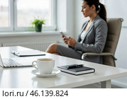 Девушка в деловом костюме работает в офисе с чашкой кофе на столе и проверяет сообщение на телефоне. Стоковое AI-изображение, AI-художник Сергей Никифоров-Гордеев / Фотобанк Лори