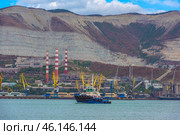 Novorossiysk port against the backdrop of the famous Markotkh ridge. In the foreground is a tugboat, and in the center are three red-and-white chimneys of the Novorossiysk cement plant, one of the oldest enterprises in the region. Numerous port cranes and warehouses are located along the shore.A tugboat in the port waters.An industrial zone with cement plant chimneys.Yellow port cranes unloading ships.The mountains of the Markotkh ridge with their characteristic limestone layers.Industrial buildings and warehouse complexes.Dense vegetation on the mountain slopes. (2023 год). Редакционное фото, фотограф Анна Кривицкая / Фотобанк Лори