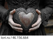 Pale female hands holding a symbolic heart made of stone surrounded by cracked black bark. The woman is dressed in a dark, antique dress with lace, giving the image a gothic atmosphere. The color scheme of the frame is maintained in gray, black and pale tones, creating a feeling of mystery, mysticism and coldness. The stone heart symbolizes emotional isolation, loss of feelings or the immutability of the internal state. Стоковое AI-изображение, AI-художник Анна Кривицкая / Фотобанк Лори