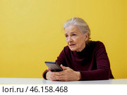 Elderly woman holding smartphone, confused expression, having phone issue, struggling with technology, sitting indoors, yellow background, facing technical challenge, thoughtful look. Стоковое AI-изображение, AI-художник Павел Муравьев / Фотобанк Лори