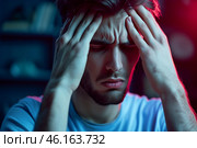 Young man expressing stress and frustration while coping with overwhelming emotions in a dimly lit room during evening hours. Стоковое AI-изображение, AI-художник Sergey Grishin / Фотобанк Лори