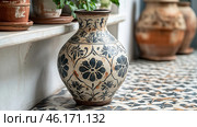 A beautiful painted ceramic vase with a black and white ornament, decorated with floral patterns. Its surface is slightly aged, which gives the product authenticity and a vintage character. The vase stands on a patterned tile, whose motifs echo the pattern on the vase itself, creating a harmonious composition. Around it you can see other terracotta pots with plants, as well as a wooden shelf with an aged effect. The atmosphere of the photo conveys the coziness of a traditional Mediterranean or oriental interior, filled with natural materials, handcrafted work and attention to detail. Стоковое AI-изображение, AI-художник Анна Кривицкая / Фотобанк Лори