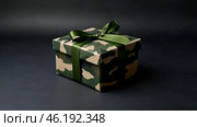 The concept of Defender of the Fatherland Day on February 23. Gift camouflage box with a festive satin ribbon bow on a dark green background. Стоковое AI-изображение, AI-художник Photojuli / Фотобанк Лори
