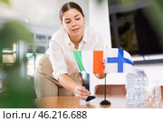 Positive young woman putting flag of Italy on the table with flag of Finland in conference room. Стоковое фото, фотограф Яков Филимонов / Фотобанк Лори