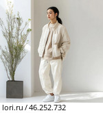 The young girl is dressed in a minimalist, modern style, with restraint and style. She wears a loose beige and milky bomber jacket, a light sweatshirt, loose white cargo pants and white sneakers. Her hair is pulled back into a low ponytail, and her expression is calm and thoughtful. The background is light, neutral, with a minimum of details, including a small green plant in a gray pot. The whole scene is designed in the aesthetics of minimalism, natural shades and urban style. Стоковое AI-изображение, AI-художник Анна Кривицкая / Фотобанк Лори