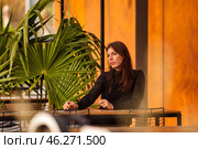A young woman sits pensively at a wooden table, surrounded by captivating orange walls and tropical plants. Sunlight filters through, casting warm hues as she reflects inwardly on her thoughts. Стоковое фото, фотограф Aleksandr Golubev / Фотобанк Лори