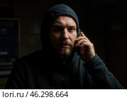 Мужчина в темном капюшоне с сосредоточенным взглядом разговаривает по телефону. Его выражение лица выдает напряженность, что может свидетельствовать о возможном мошенничестве. Стоковое AI-изображение, AI-художник Сергей Никифоров-Гордеев / Фотобанк Лори