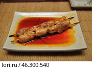 Two skewers of chicken shashlik cooked in a glaze or sauce, served on a square plate. The sauce has a rich red-orange color and has spread across the plate, giving the dish an appetizing look. The background is a bamboo mat, creating an oriental style of serving. Perhaps this is a Japanese dish yakitori or another Asian dish on skewers. Стоковое фото, фотограф Анна Кривицкая / Фотобанк Лори