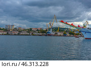 Industrial part of Sevastopol Bay, where numerous port cranes, shipyards and docks are visible. On the right side of the frame, a beautiful sailboat with a decorated bow is partially shown, which adds to the contrast between the modern shipbuilding industry and the maritime romance.The urban development in the background combines old buildings of historical value with modern high-rise buildings. Thick clouds over the bay add drama to the scene and emphasize the harsh nature of the seaside city. The water is calm, with slight ripples reflecting the sky. (2021 год). Редакционное фото, фотограф Анна Кривицкая / Фотобанк Лори