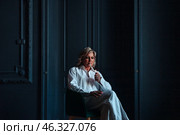 A middle-aged woman sits comfortably in a modern indoor setting, lost in thought while elegantly holding a object. Soft shadows and rich textures surround her, creating an atmosphere of tranquility. Стоковое фото, фотограф Aleksandr Golubev / Фотобанк Лори