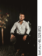 A well-dressed man sits in a plush chair, wearing a crisp white shirt and stylish hat. His composed demeanor creates an atmosphere of elegance in a tastefully decorated indoor space. Стоковое фото, фотограф Aleksandr Golubev / Фотобанк Лори