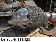Medieval knight's helmet - bascinet with aventail (chainmail protection for the neck). The helmet is made of metal, polished to a shine, but has traces of scratches and abrasions, which indicates its active use, possibly in historical reconstructions or buhurts. Breathing holes are visible, as well as chainmail protecting the warrior's neck.Nearby are elements of armor: gauntlets, parts of bracers and fragments of a shield. The helmet with a pointed shape was popular among the knights of Europe in the 14th-15th centuries and was used in battle along with a cuirass and plate armor. Стоковое фото, фотограф Анна Кривицкая / Фотобанк Лори