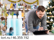 Bearded man seller restock of goods using tablet in store decorated for New Year holiday. Стоковое фото, фотограф Яков Филимонов / Фотобанк Лори