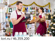 Focused bearded man seller restock of goods using tablet, female assistant laying out goods in background in store decorated for New Year holiday. Стоковое фото, фотограф Яков Филимонов / Фотобанк Лори