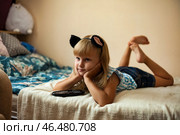 A cheerful child lies comfortably on a bed, resting her chin on her hands. The playful atmosphere is enriched by her cat-ear headband, inviting imagination and warmth. Стоковое фото, фотограф Aleksandr Golubev / Фотобанк Лори