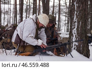 Sestroretsk, Russia - February 19, 2023: Two soldiers huddle in the snow, repairing their weapon as winter's chill descends, surrounded by tall trees and signs of battle. Редакционное фото, фотограф Aleksandr Golubev / Фотобанк Лори