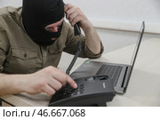 Мошенник в черной балаклаве у ноутбука с телефонной трубкой в руке. Стоковое фото, фотограф Сергей Булкин/NEWS.ru / ТАСС / Фотобанк Лори