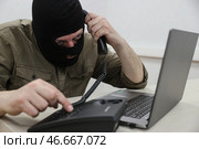 Мошенник в черной балаклаве у ноутбука с телефонной трубкой в руке. Стоковое фото, фотограф Сергей Булкин/NEWS.ru / ТАСС / Фотобанк Лори