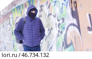 Serious young man in casual outfit and balaclava walking along the grafitti wall. Стоковое фото, фотограф David Kashakhi / Фотобанк Лори