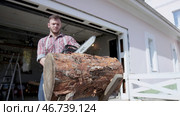 Brutal young man cuts piece of log with saw on background of garage. Стоковое фото, фотограф David Kashakhi / Фотобанк Лори