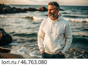 A handsome middle-aged man with a beard wearing a white hoodie on the beach. Template layout for hoodie design, print area layout for logo or design. Стоковое AI-изображение, AI-художник Photojuli / Фотобанк Лори