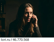 Молодая женщина разговаривает по проводному телефону, возможно, став жертвой обмана в темном офисе. Стоковое AI-изображение, AI-художник Сергей Никифоров-Гордеев / Фотобанк Лори