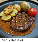 A delicious steak with characteristic grill marks, served with a side of baked potatoes and tomatoes. The dish is garnished with a sprig of fresh thyme and topped with a thick, peppery meat sauce. The potatoes are browned and the tomatoes are lightly roasted, giving the composition rich colors. The black plate creates a contrast, highlighting the rich shades of the meat and side dish. The atmosphere of the dish speaks of a premium restaurant or haute cuisine. Стоковое AI-изображение, AI-художник Анна Кривицкая / Фотобанк Лори