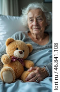In a quiet hospital room, an elderly woman sits calmly with a teddy bear in her lap, finding solace and companionship while confronting her hardships and loneliness. Стоковое AI-изображение, AI-художник Юлия Петровская / Фотобанк Лори