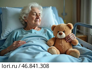 An elderly woman rests in a hospital bed, eyes peacefully closed. Beside her, a soft teddy bear symbolizes comfort and companionship, providing warmth in her moments of hardship. Стоковое AI-изображение, AI-художник Юлия Петровская / Фотобанк Лори